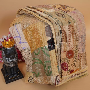 Puede incluir: Una colcha kantha beige con un dise&ntilde;o de patchwork que presenta varios patrones florales y geom&eacute;tricos. La colcha est&aacute; hecha de saris reciclados y est&aacute; cosida a mano con bordado kantha. La colcha est&aacute; doblada y expuesta sobre una mesa con una peque&ntilde;a estatua de le&oacute;n en primer plano.