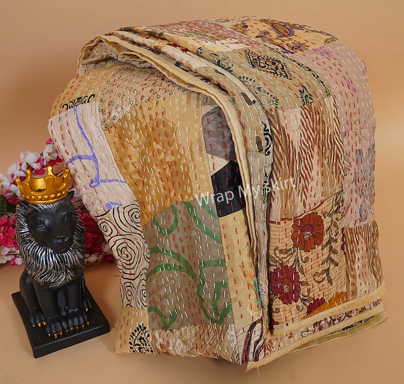 Puede incluir: Una colcha kantha beige con un dise&ntilde;o de patchwork que presenta varios patrones florales y geom&eacute;tricos. La colcha est&aacute; hecha de saris reciclados y est&aacute; cosida a mano con bordado kantha. La colcha est&aacute; doblada y expuesta sobre una mesa con una peque&ntilde;a estatua de le&oacute;n en primer plano.