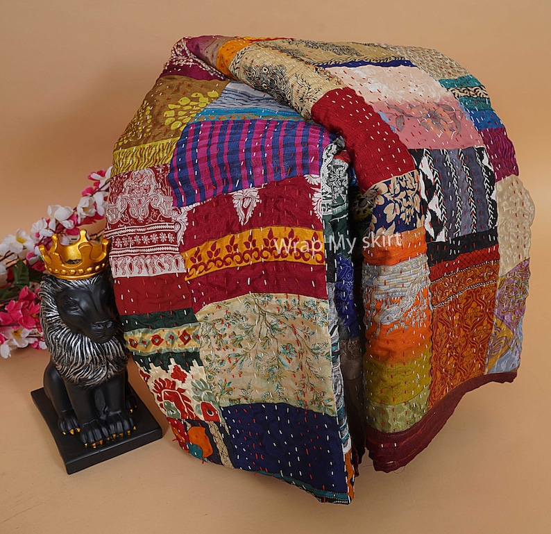 Pu&ograve; includere: Una trapunta colorata a patchwork realizzata con sari riciclati. La trapunta &egrave; composta da quadrati di tessuti, colori e motivi diversi. La trapunta &egrave; piegata a met&agrave; e si trova su una superficie marrone chiaro.