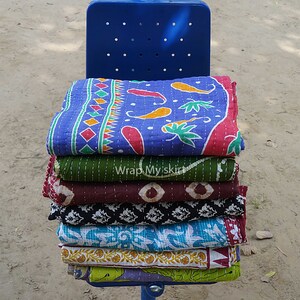 May include: A stack of colourful kantha quilts with various patterns, including floral, geometric, and paisley designs. The quilts are folded and stacked on a blue metal bench.