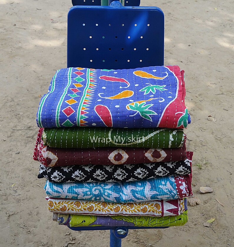 May include: A stack of colourful kantha quilts with various patterns, including floral, geometric, and paisley designs. The quilts are folded and stacked on a blue metal bench.