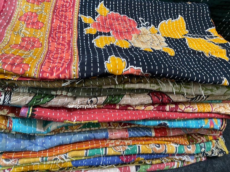 Op de afbeelding: Een stapel kleurrijke kantha-dekens met bloemenpatronen. De dekens zijn gemaakt van gerecyclede sari's en zijn met de hand gestikt. De bovenste deken is zwart met gele en rode bloemmotieven.