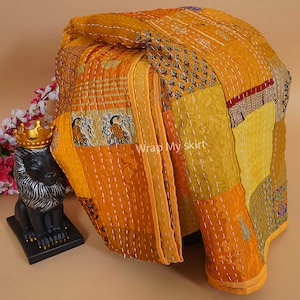 Pu&ograve; includere: Una trapunta kantha gialla e dorata con un design a patchwork. La trapunta &egrave; realizzata con sari riciclati ed &egrave; cucita a mano. La trapunta &egrave; piegata ed esposta su un tavolo.