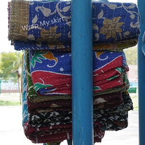 May include: A stack of colourful kantha quilts with floral and geometric patterns. The quilts are folded and stacked on a blue metal pole. The text "Wrap My Skirt" is visible on the top quilt.