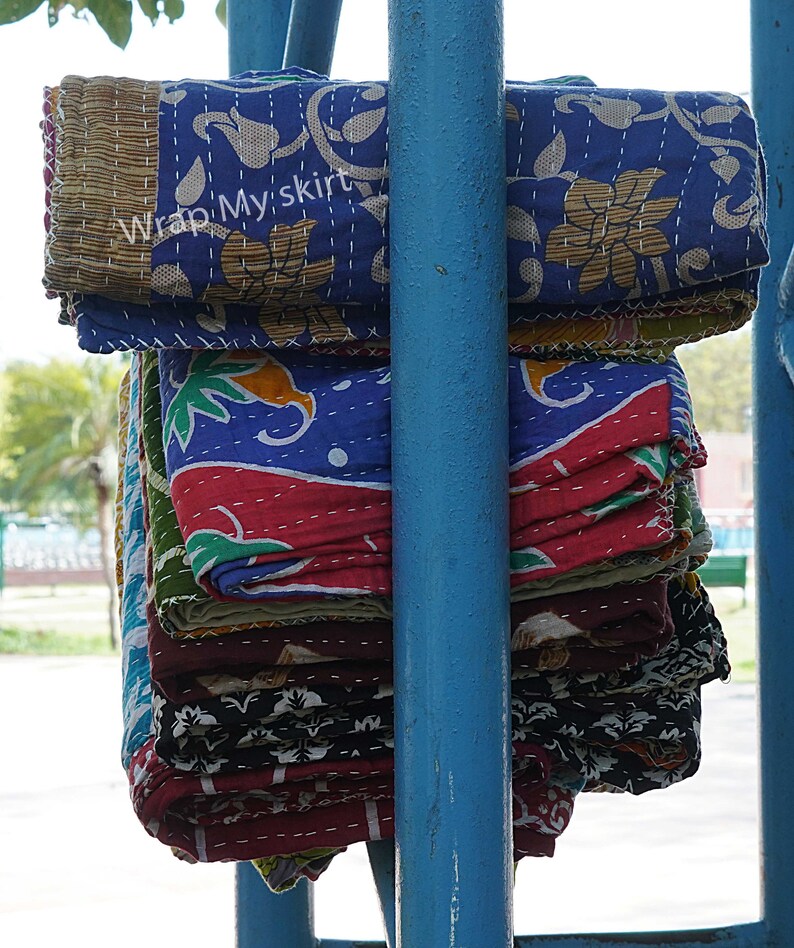 May include: A stack of colourful kantha quilts with floral and geometric patterns. The quilts are folded and stacked on a blue metal pole. The text "Wrap My Skirt" is visible on the top quilt.