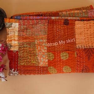 以下が含まれることがあります： オレンジ、ブラウン、クリームの色合いの様々な生地で作られたパッチワークキルト。キルトは白い糸で手縫いされており、独特の素朴な外観をしています。キルトには「Wrap My skirt」という文字が見えます。