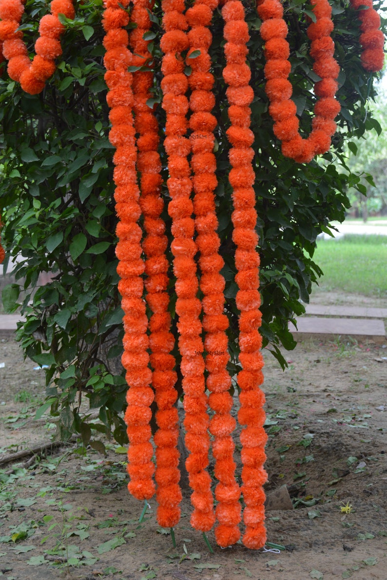 May include: A garland of orange marigold flower pom-poms hanging from a tree branch. The garland is made of multiple strands of flowers, each strand is about 90 cm long.