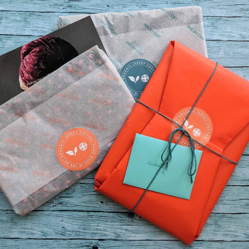 Gift wrapped hair towel in bright orange recyclable wrapping paper and branded sticker in orange, tied with teal coloured string. Teal envelope containing gift card. Towels scattered around wrapped in branded tissue paper secured with stickers.
