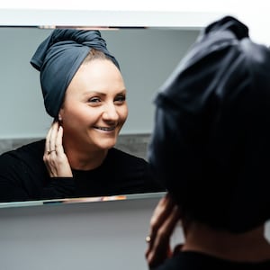 A woman wearing a dark grey organic jersey good wash day towel on her head, looking into a mirror.