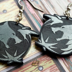 Puede incluir: Un par de pendientes plateados y negros con el logotipo de The Witcher, una cabeza de lobo dentro de un círculo con una huella de pata de lobo estilizada.