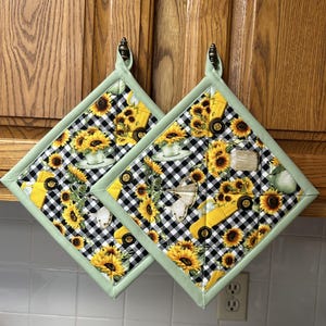 May include: Two square pot holders with a black and white checkered pattern, featuring yellow sunflowers, a yellow truck, and a teacup. The pot holders have a light green border and a loop for hanging.