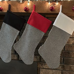May include: Three Christmas stockings hang from a wooden mantel. Each stocking has a black and white checkered pattern on the body. The cuffs are solid black, red, and white. Red ornaments and string lights decorate the mantel.