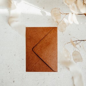 May include: A brown paper envelope on a white textured background with a dried flower branch in the upper right corner.