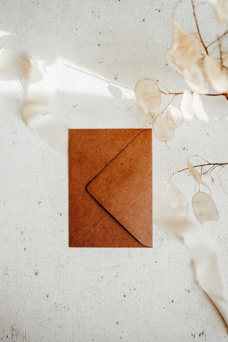 May include: A brown paper envelope on a white textured background with a dried flower branch in the upper right corner.