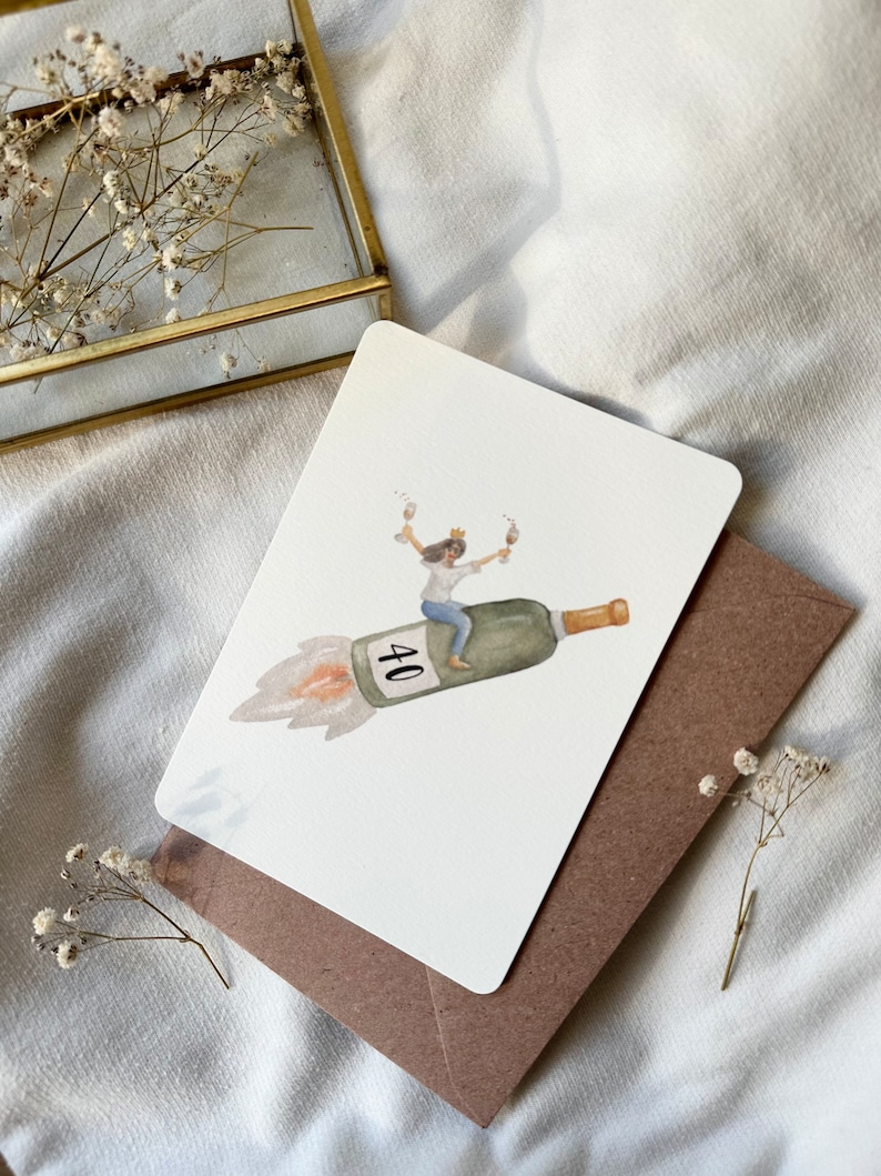 May include: A white card with a watercolor illustration of a person wearing a crown and riding a champagne bottle shaped like a rocket. The bottle is labeled with the number "40".