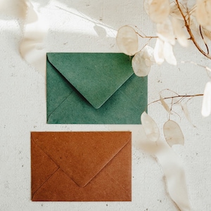 May include: Two envelopes, one green and one brown, are lying on a white textured surface. A white ribbon and dried flowers are in the background.