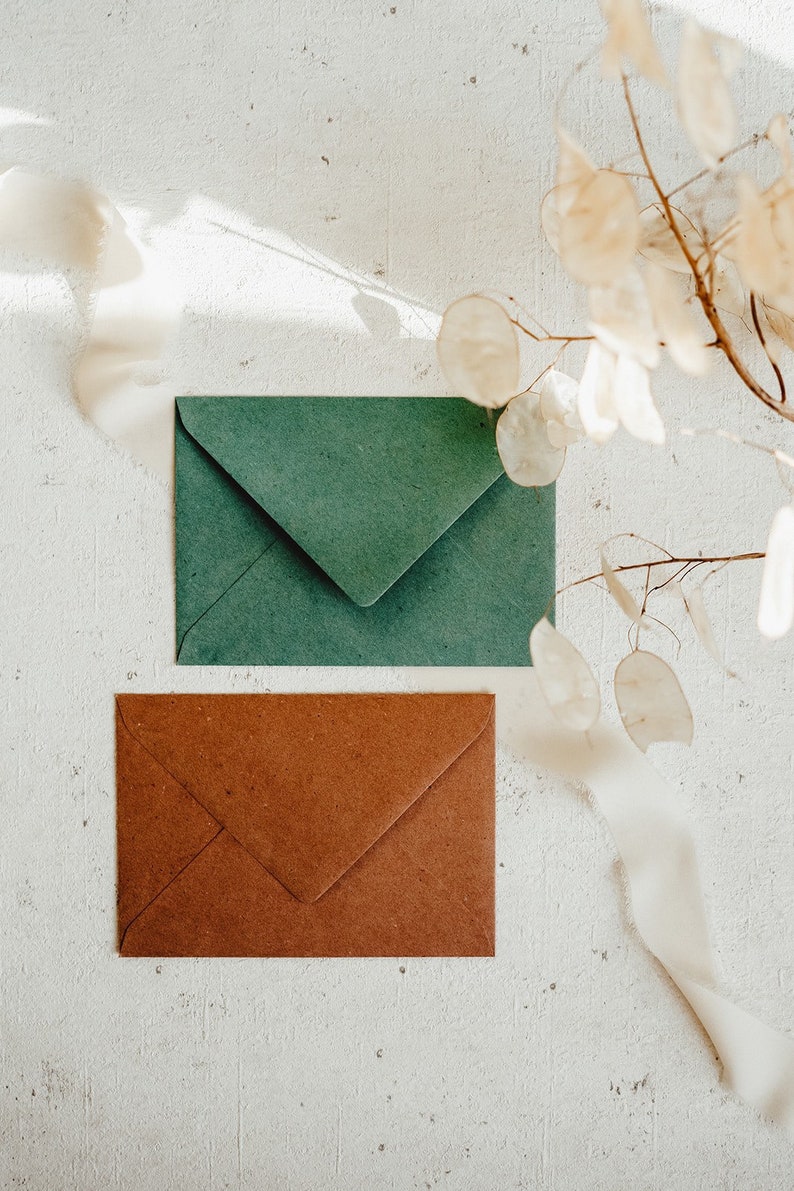 May include: Two envelopes, one green and one brown, are lying on a white textured surface. A white ribbon and dried flowers are in the background.