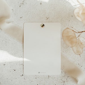 May include: A blank white card with a small round sticker in the top center that says "Linda Mundo". The card is on a white textured surface with a white ribbon and dried flowers.