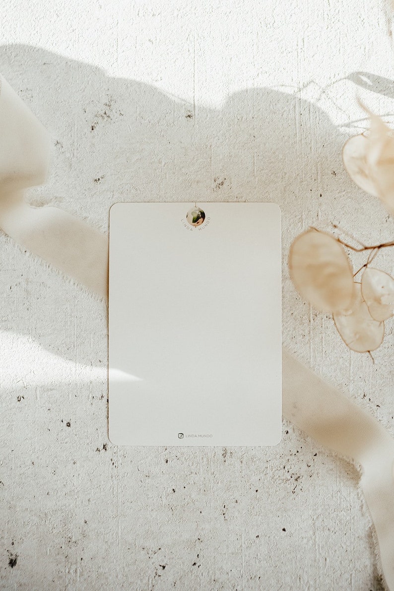 May include: A blank white card with a small round sticker in the top center that says "Linda Mundo". The card is on a white textured surface with a white ribbon and dried flowers.