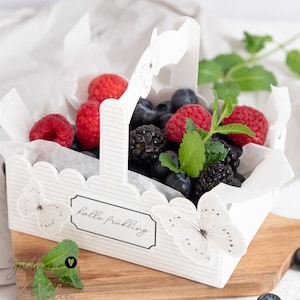 Könnte beinhalten: Ein weißer Papierkorb mit Henkel, verziert mit Schmetterlingen, gefüllt mit Blaubeeren, Himbeeren und Brombeeren. Der Korb hat ein Etikett mit der Aufschrift "hallo fruehling".