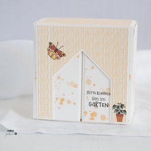 Może przedstawiać: Biała kartka z żółtym paskowanym tłem i ilustracją motyla. Kartka jest złożona w kształt domu z tekstem "Bitte Klingeln Bin im Garten" na przodzie.