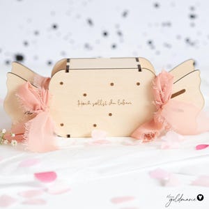 Puede incluir: Una caja de madera con forma de caramelo con una ranura para insertar dinero. La caja es de color marrón claro con puntos marrones oscuros y el texto "Hoch sollst du leben" grabado en la parte delantera. Cintas rosas y pétalos de flores añaden un toque decorativo.