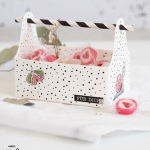 Könnte beinhalten: Weiße Geschenkbox mit schwarzen Punkten und einem schwarz-weiß gestreiften Griff. Die Box hat drei Fächer, die mit rosa herzförmigen Süßigkeiten gefüllt sind. Die Box hat ein Etikett mit der Aufschrift "FÜR DICH", was auf Deutsch "für dich" bedeutet.