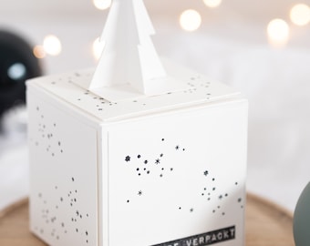 May include: A white gift box with a pop-up paper Christmas tree on top. The box has a black and white star pattern and the text "MIT LIEBE VERPACKT" on the front.