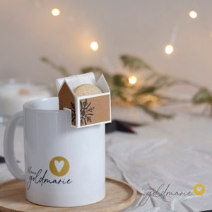 Può includere: Una tazza in ceramica bianca con un cuore dorato e il testo "Liebe goldmarie" sul lato. Una piccola scatola di carta marrone con un motivo a fiocco di neve contiene un biscotto ed è attaccata alla tazza.