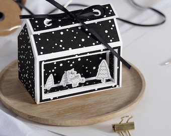 May include: A black and white paper house with a black ribbon tied around it. The house has a window with a white background and black polka dots. Inside the window are three paper cutouts of a small tree and a bunny wearing a scarf. The house is sitting on a wooden tray.
