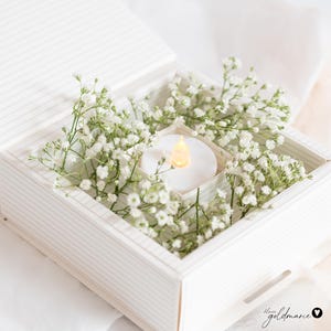 Può includere: Una scatola bianca a righe piena di delicati fiori bianchi di gipsofila e una piccola candela bianca rotonda con una luce gialla tremolante. L'insieme crea un'atmosfera morbida e romantica.