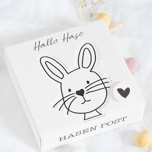 May include: A white box with a black and white illustration of a bunny rabbit. The box has the text "Hallo Hase" and "Hasen Post" on it.