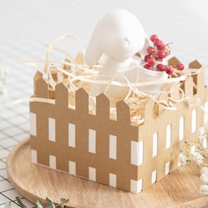 May include: A white ceramic bunny figurine sits inside a brown cardboard box with a picket fence design. The box is filled with shredded paper and red berries.