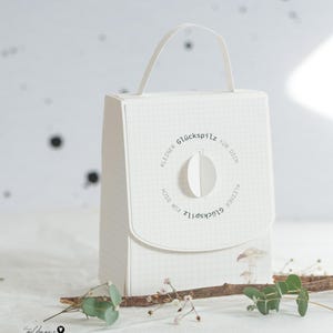 以下が含まれることがあります： ハンドル付きの白い紙製のギフトバッグ。格子模様と円形のフラップが特徴です。 フラップには、ドイツ語のテキスト「Kleiner Glückspilz für Dich」が円形に配置されています。 バッグは小枝と小さな植物で飾られています。