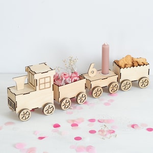 Puede incluir: Un tren de juguete de madera con cuatro vagones, decorado con confeti rosa y blanco. El tren tiene el número "6" en el segundo vagón, un pequeño ramo de flores blancas en el tercer vagón y caramelos rosas en el cuarto vagón. El último vagón tiene una vela marrón en la parte superior.