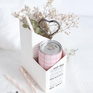 Op de afbeelding: Een witte kartonnen doos met een roze-wit gestreepte blikje erin. De doos heeft een hartvormige uitsparing en is versierd met een citaat over vriendschap, lachen en avontuur. De doos is gevuld met witte bloemen en roze-wit gestreepte rietjes.