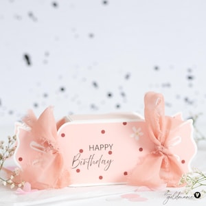 Puede incluir: Una caja de regalo rosa con el mensaje "Happy Birthday", adornada con lunares rojos y una flor blanca. La caja está atada con una delicada cinta color melocotón. El fondo es blanco con confeti negro disperso.