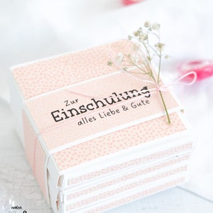 May include: A stack of pink gift boxes with white polka dots and a white ribbon tied around the top box. The top box has the text "Zur Einschulung alles Liebe & Gute" printed on it.