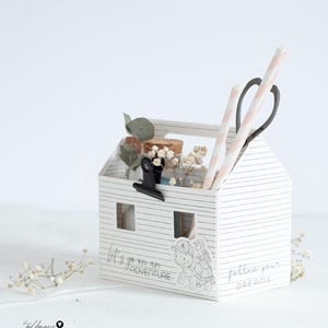 Könnte beinhalten: Ein weißer, hausförmiger Organizer mit horizontalen Streifen, der dekorative Gegenstände enthält. Er zeigt den Text "let's go on an adventure" und "follow your dreams". Der Organizer enthält Strohhalme, eine Schere und kleine Gläser mit Blumen.