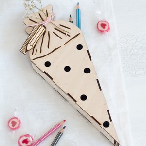 Könnte beinhalten: Ein Holz-Bleistifthalter in Form eines Kegels mit einem ausgeschnittenen Design einer Tasche. Der Halter hat Löcher für Bleistifte und ist mit schwarzen Punkten verziert. Es gibt drei Bleistifte und vier rosafarbene herzförmige Bonbons auf einer weißen Oberfläche.