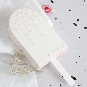 Puede incluir: Caja blanca en forma de paleta de helado con lunares rosas y un diseño de goteo rosa. La caja tiene un palo blanco y dice "Lass dich feiern" en texto negro.