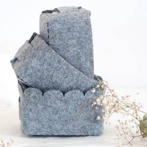 Puede incluir: Tres cestas de almacenamiento apiladas de fieltro gris. La cesta superior es rectangular, la del medio está en ángulo y la inferior tiene un borde festoneado. Las cestas están hechas de un material suave y texturizado.
