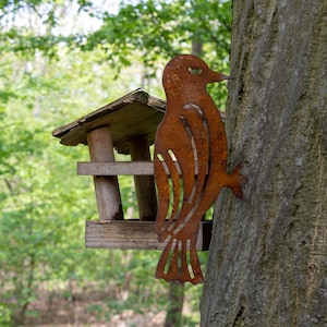 May include: A wooden bird feeder with a rusty metal bird silhouette attached to a tree trunk. The feeder has a small roof and two perches.