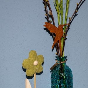 May include: A green fabric flower with a white button centre on a wooden stick with white leaves. A blue glass vase with a textured surface holds a bouquet of green branches with a rusty metal hummingbird decoration.