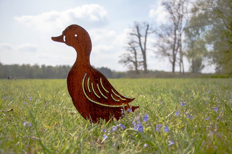 Peut inclure: Une silhouette de canard en m&eacute;tal rouill&eacute; debout dans un champ d'herbe verte avec des fleurs sauvages bleues.