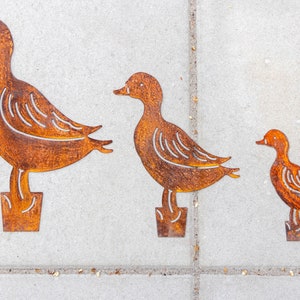 Peut inclure: Trois silhouettes de canards en m&eacute;tal de diff&eacute;rentes tailles, en m&eacute;tal brun rouill&eacute;, debout sur une surface en b&eacute;ton gris.