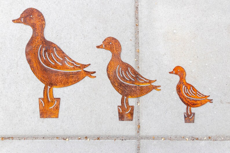 Peut inclure: Trois silhouettes de canards en m&eacute;tal de diff&eacute;rentes tailles, en m&eacute;tal brun rouill&eacute;, debout sur une surface en b&eacute;ton gris.