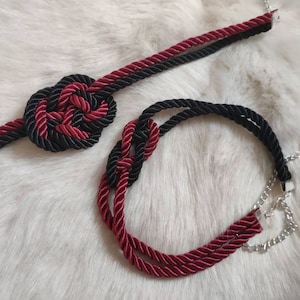 Puede incluir: Dos collares de cuerda trenzada, uno con un nudo en forma de corazón en negro y burdeos, el otro con un acento de cadena plateada.