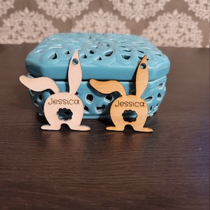 Puede incluir: Dos etiquetas de Pascua de madera en forma de conejo con el nombre "Jessica" escrito en cada una. Las etiquetas están sobre una superficie de madera marrón oscuro delante de una caja decorativa azul y blanca.