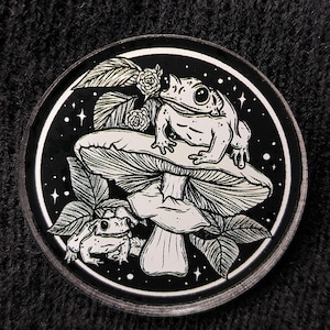 May include: A black and white enamel pin featuring a frog sitting on a mushroom with a smaller frog below. The pin has a starry night background with leaves and flowers.
